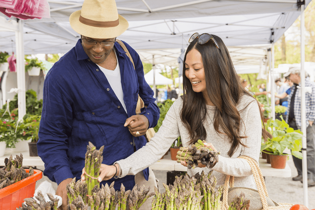 Man in hat and blue shirt and brunette woman shopping and open-air market buying fresh Asparagus under white tent.